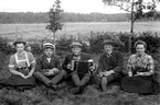 TRE MÄN OCH TVÅ KVINNOR SITTER I GRÄSET. MANNEN SOM SITTER I MITTEN SPELAR DRAGSPEL.