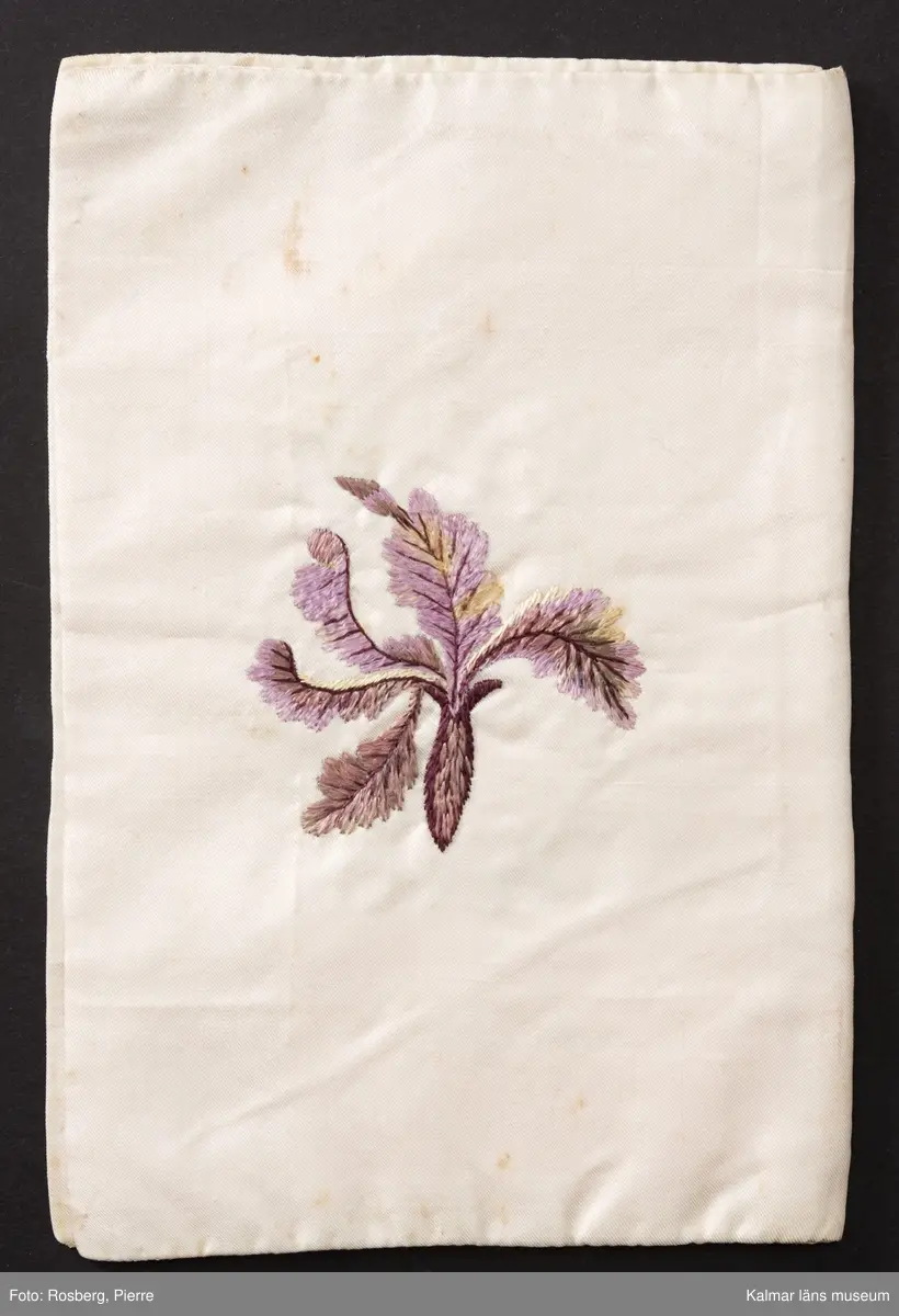 KLM 8909. Sedelbok, plånbok. Av vitt siden med broderier av schattérsöm, plattsöm, knutsöm, kjedjesöm. På in- och utsidornas fält blomdekorer. Insidorna märkta inom blomsterkransar med text: M G F 1818. Bröllopsgåva, fästegåva till Magnus Gudmundsson Fröberg.