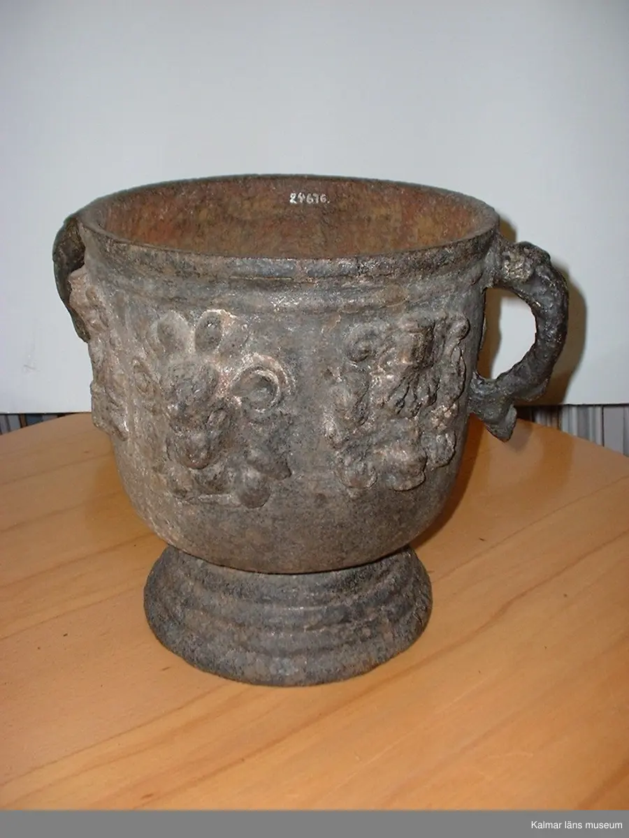 KLM 24676. Trädgårdsurna med fot, av järn, gjutjärn. Med två öron i form av delfiner. Dekor i relief med vädurshuvuden och adligt vapen (icke identifierat), växelvis. Ett av vapnen vänt upp och ner. I urnans botten 4 små runda hål för regnvatten att rina ut genom. Datering, 1600-talets mitt.