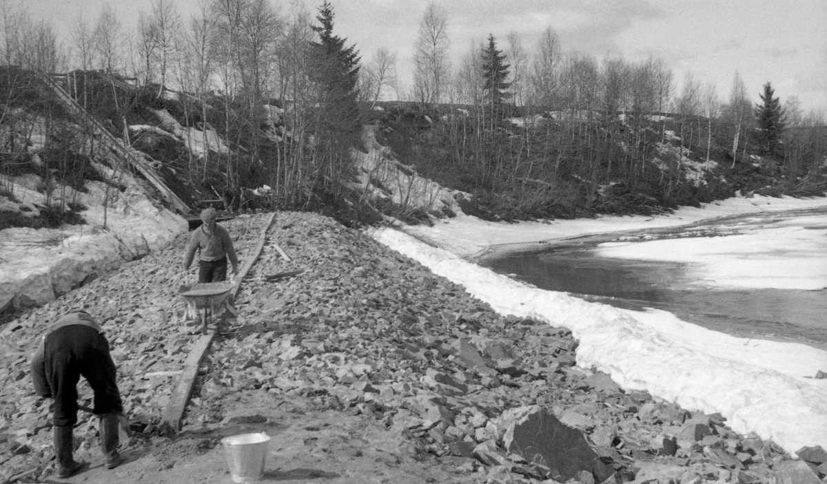 Bygging av skådam i en elvesving i Glomma i nærheten av Kalvskjæret ved Braskereidfoss i Våler i Solør. Dammen var en steinvoll som tilsynelatende skulle gis et ytre skall av sement. Mørtelen ble tilsynelatende blandet i et kar i den andre enden av vollen og deretter kjørt ut med trillebår på ei rekke av planker som var lagt etter hveradre langs midten av vollen. I forgrunnen ser vi en mann som jevnet ut mørtelen ved hjelp av en spade. I den bratte elveskråningen i bakgrunnen var det ei renne, antakelig for materialer som skulle brukes under dambygginga. Bildet ble tatt våren 1946.