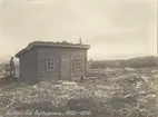 Första stugan vid Sylarna. Litet hus byggt av liggande timmer. Snedtak, två fönster och en dörr. En man i hatt står bakom stugan. Nyröjd mark runt huset, siluetter av fjäll i bakgrunden.