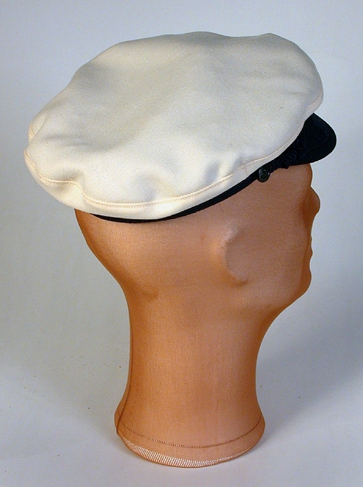 Vit herrmössa med platt kulle. Snodd mitt fram som knäppes i sidorna. 
   Mått: Runt kullen 620 mm. Kullens höjd 80 mm. 
   Storlek: 56.