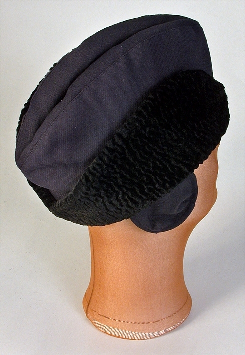 Herrmössa, båtformad av blått ylletyg. Söm mitt fram och mitt bak. Uppvikt brätte klätt med krimmer. Öronlappar som är nedfällbara.