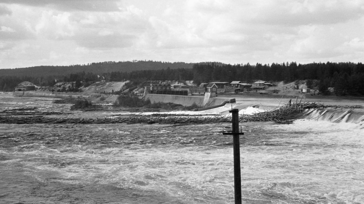 Skjefstadfossen i Glomma, nærmere bestemt i Heradsbygda i Elverum, Hedmark, fotografert i slutten av mai 1947. Her hadde man i 1909-1910 bygd et elvekraftverk med en terskeldam som skulle stuve opp vann til turbinene i en kraftstasjon som ble reist ved vestre elvebredd, nederst ved fossen (til venstre på dette fotografiet). Overløpsdammen var 312 meter lang, utført i betong, støpt i bue med den konvekse sida mot strømretningen og slik at damkrona var noe lavere i den østre enn i den vestre enden. Hensikten var å skape et «sug» i overflata som trakk fløtingstømmeret mot den østre delen av elveløpet. Den nedenforliggende fossen hadde nemlig et bredt, grunt og steinete løp der tømmeret hadde lett for å binde seg og danne hauger. Akkurat det hadde øyensynlig skjedd da dette fotografiet ble tatt. Vi ser en langstrakt tømmerhaug, som må ha oppstått ved at noen stokker hektet seg fast i oppstikkende steiner nord for Tjuvholmen, som delte elveløpet i to. De første stokkene som satte seg fast her fanget løstømmer som kom flytende ovenfra, og slik bygde tømmerhaugen seg oppover mot fallet fra overløpsdammen. Her ble det en arbeidskrevende og ikke helt ufarlig oppgave for fløterne arbeide stokkene ut i de strømmende vannmassene igjen. Til høyre i bildet ser vi Skjefstadfossdammens inntaksbasseng med sine lukehus. Vanninntaket til kraftstasjonen lå ved det vestre landfestet, der det i 1909 ble støpt et drøyt 400 meter langt betongrør nedover mot kraftstasjonen. I 1916 ble det støpt ytterligere et slikt rør, parallelt med det første. Kraftstasjonen ble bygd i teglmur og betong. Den inneholdt fra starten av tre turbiner, men disse ble supplert med ytterligere to, i 1918 og 1939.