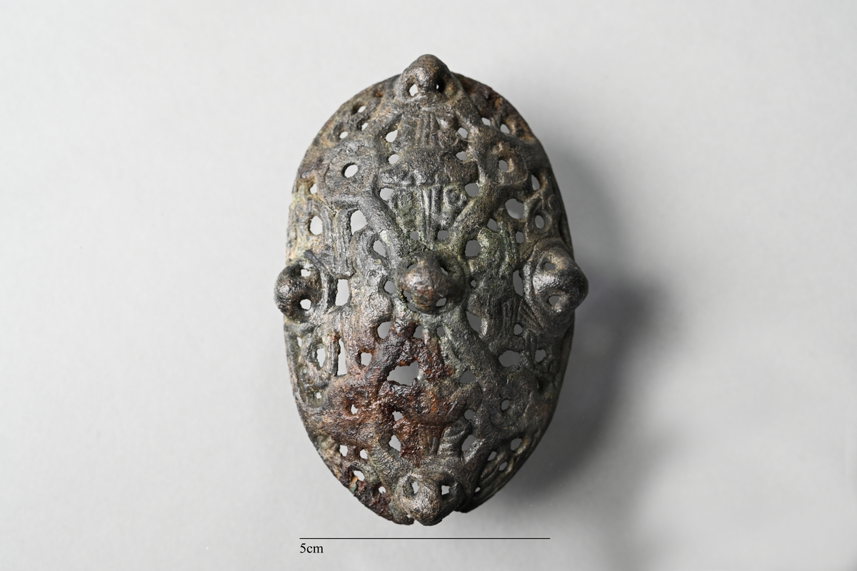 Spännbuckla (se Petersen fig 51) med genombruten överskålla med fem knoppar. Överskållan hel, separerad från underskållan. Underskållan skadad, ornerad längs kanten. Nålfäste och nålhållare kvar, men ej nålen. Tygavtryckmärken från gjurtformen på undersidan, troligen från linne vävt i tuskaft.
