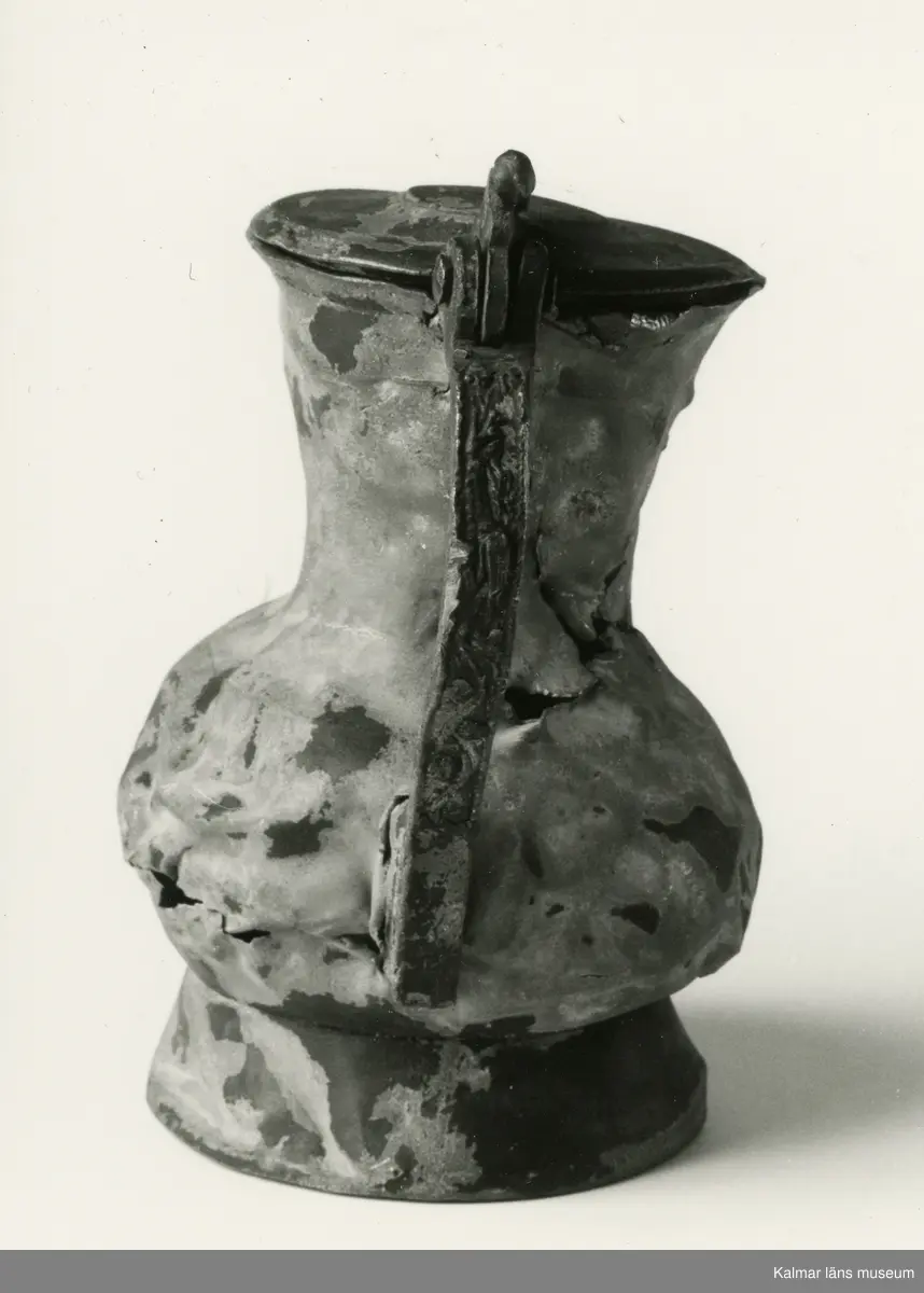 KLM 46428 Kanna av tenn. Bukig form. Hankkärl, försedd med lock och handtag, vidgad botten neråt. I botten, invändigt en medalj med korsfästelsebild. Medaljens kant försedd med pärlring. Ägarmärke/bomärke på lockets och bottens utsidor. Handtaget ornerat med drakmotiv och bladslinga, tumgreppet saknar ett av ekollonen. Kannan skadades svårt vid upphittandet, genomgående sprickor på många ställen.