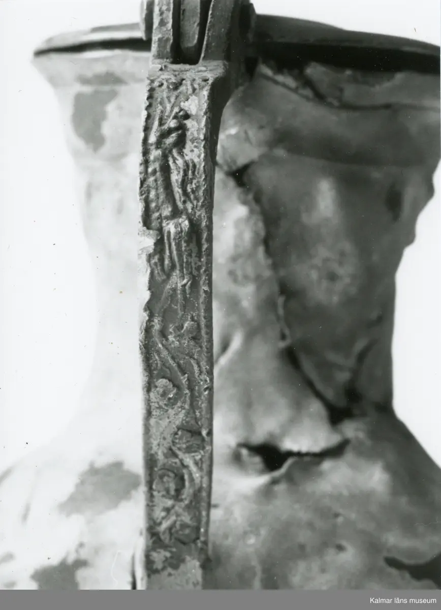 KLM 46428 Kanna av tenn. Bukig form. Hankkärl, försedd med lock och handtag, vidgad botten neråt. I botten, invändigt en medalj med korsfästelsebild. Medaljens kant försedd med pärlring. Ägarmärke/bomärke på lockets och bottens utsidor. Handtaget ornerat med drakmotiv och bladslinga, tumgreppet saknar ett av ekollonen. Kannan skadades svårt vid upphittandet, genomgående sprickor på många ställen.