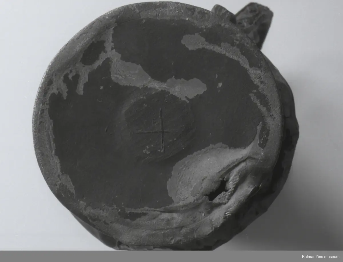 KLM 46428 Kanna av tenn. Bukig form. Hankkärl, försedd med lock och handtag, vidgad botten neråt. I botten, invändigt en medalj med korsfästelsebild. Medaljens kant försedd med pärlring. Ägarmärke/bomärke på lockets och bottens utsidor. Handtaget ornerat med drakmotiv och bladslinga, tumgreppet saknar ett av ekollonen. Kannan skadades svårt vid upphittandet, genomgående sprickor på många ställen.