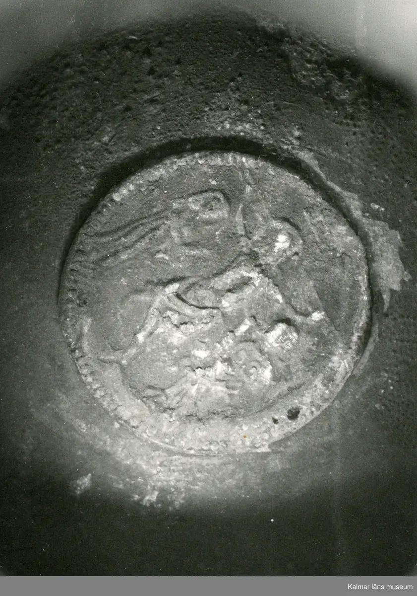 KLM 46428 Kanna av tenn. Bukig form. Hankkärl, försedd med lock och handtag, vidgad botten neråt. I botten, invändigt en medalj med korsfästelsebild. Medaljens kant försedd med pärlring. Ägarmärke/bomärke på lockets och bottens utsidor. Handtaget ornerat med drakmotiv och bladslinga, tumgreppet saknar ett av ekollonen. Kannan skadades svårt vid upphittandet, genomgående sprickor på många ställen.