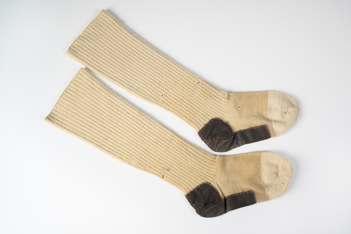 Långstrumpor till man, ett par, av beige yllegarn. Tådelen är strumporna är stickade i ljusare garn. Här och underdel av strumpan är lagad med ett smårutigt bomullstyg svart och brunt.