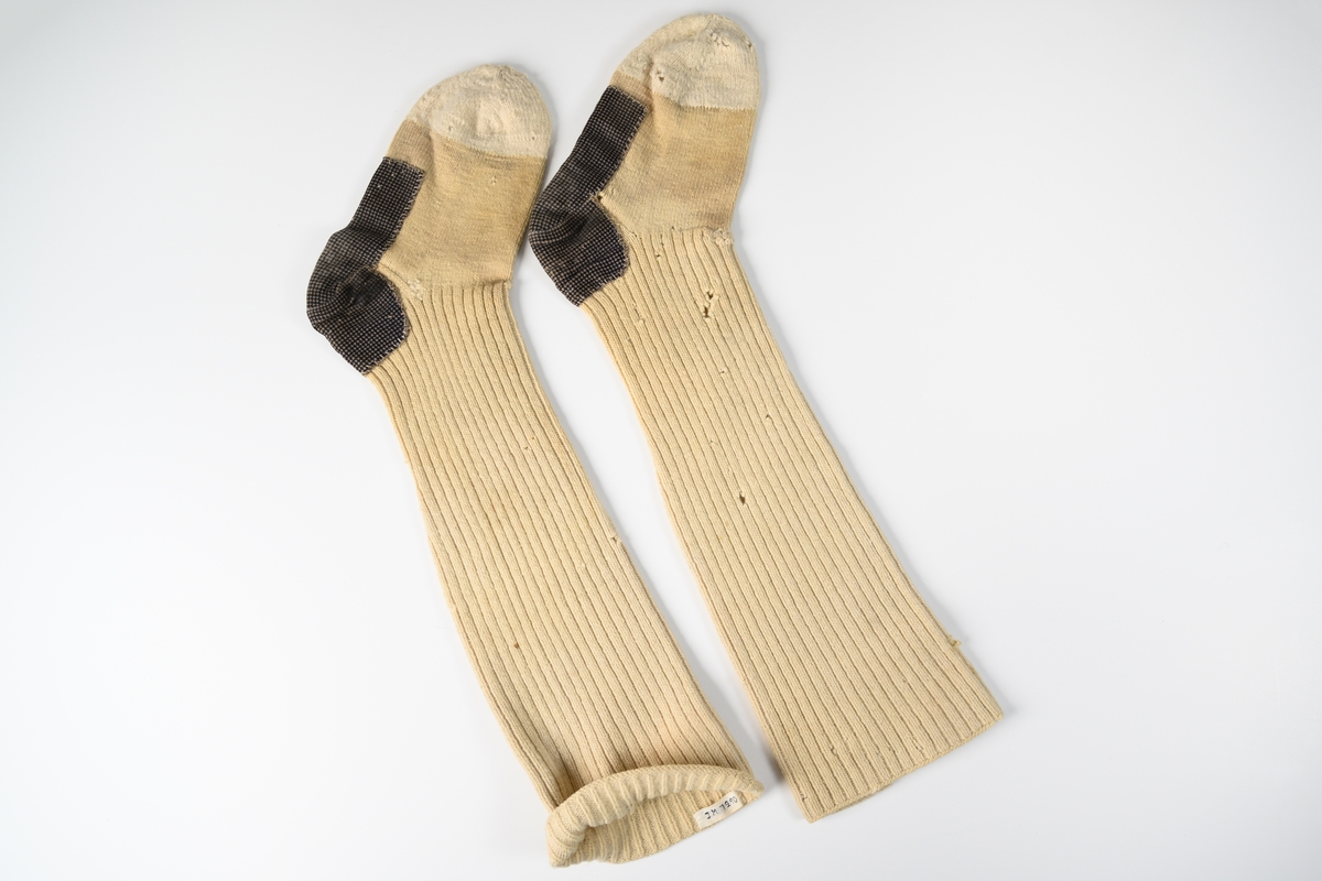 Långstrumpor till man, ett par, av beige yllegarn. Tådelen är strumporna är stickade i ljusare garn. Här och underdel av strumpan är lagad med ett smårutigt bomullstyg svart och brunt.