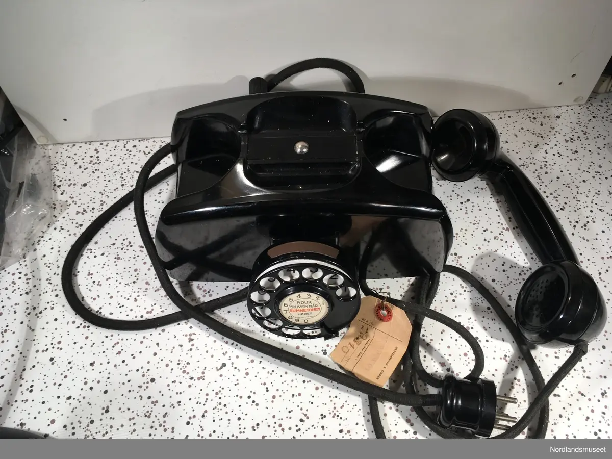 Telefonapparat med sveiv og nummerskive
Kombimodell, som kan benyttes i manuelt og automatisk telefonnett.
Bunnplate merket med AD 51775
Merkelapp på ledning merket med: "2. nov 1951" og "MEURICE BRUXELLES"
Produsent er noe usikker.