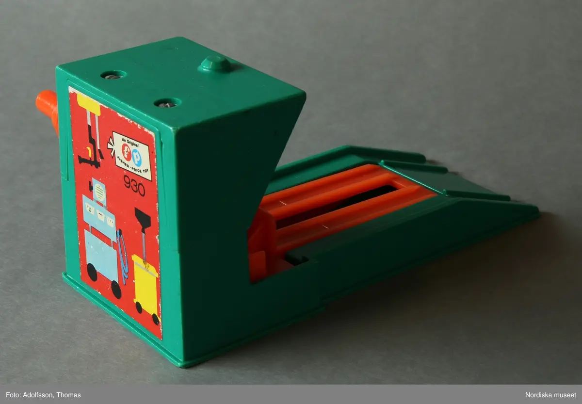 a) Garage med ramp och parkeringsplatser i två våningar för leksaksbilar. Formgjuten plast i klara färger, med påklistrade pappersetiketter med text och bild. Enkel mekanik för att höja eller sänka en hiss för tillhörande leksaksbilar med människofigurer samt roterande platta på det övre planet. Garaget står på en platta av masonit. På nedre plan finns en fast bensinpump med slang av textilt material samt b) en lös ramp med mekanik för höjning och sänkning av leksaksbilar. Till garaget hör fyra bilar som kan rulla nedför en ramp från parkeringsplatserna på det översta planet.