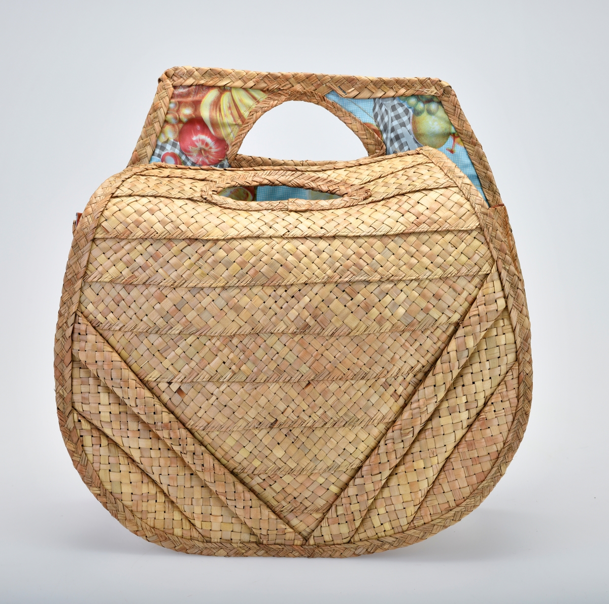 En veske av bast og plast som er flettet og maskinsydd. Basten er flettet. I front (på begge sider) er det 10 vannrette striper som er omtrent 4cm brede. Stripene blir kortere fra øverst og ned mot bunnen (den nederste stripen er kun en liten trekant). På hver side er det 3 skråstilte striper som er flettet med mer "volum". Det er trekantformede håndtak øverst på begge sider. Sidene og bunnen på vesken består av 4 flettede striper. Vesken er foret med plast. Den er blå- og hvitrutet med bilder av diverse frukt og en primus på rutete duk. Mønsteret er repeterende.