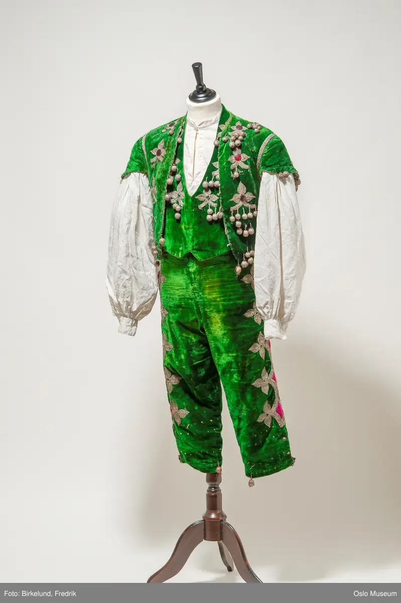 A: vest, grønn bunn med broderte blomster i metallfarget tråd. Pyntet med rød plaststeiner. 
B: bukse, grønn bunn med blomster brodert i metallfarget tråd langs kanten. Rosa dekor. 
C: skjorte, hvit lin. 
D: løse deler
E: vest