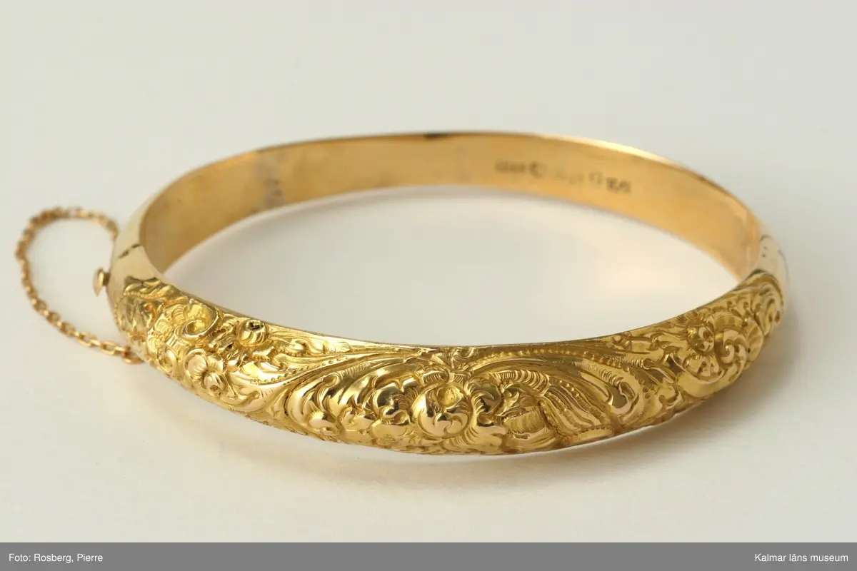 KLM 34781 Armring av guld. Oval form. Ihålig armring, slät undersida, ovansidan rikt dekorerad med blad och blommönster. Förvaras i fyrkantig pappask, gulfärgad, tillsammans med gul hårlock. Stämplar: 18K, kontrollstämpel, mästarstämpel, Giron & Löngren (1820-1847), stadsstämpel, Stockholm, årsstämpel, K4 =1840. Inskription: d 15 juni och d 9 juli 1840.