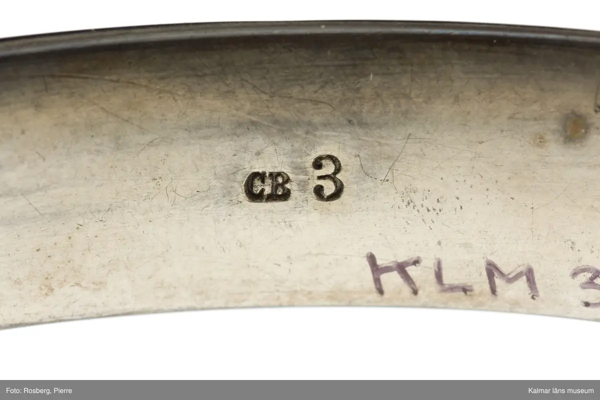 KLM 34780:1  Armring, av svart material. Oval, inläggningar av silver, infattningar av gul metall och pärlor. Förvaras i oval röd ask med text: A Michelsen, Hof og Ordens- Juveler, Kongens Nytorv. Har troligen tillhört Jenny Nyström.