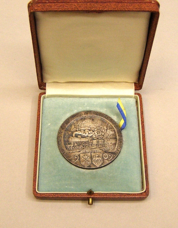 Minnesmynt av silver. På framsidan syns ett tåg draget av ett ånglok i full fart genom ett landskap, samt texten "Till minne af Gefle-Dala järnvägs femtioårsdag 13 juli 1909".
På baksidan avbildas en grankvist med kottar, samt ett vinghjul och texten "Bryta väg var af forno svensk mans största ära".
Myntet är signerat Sven Kulle på båda sidor.

Plaketten ligger i en brun ask fodrad med ljus turkos sammet.