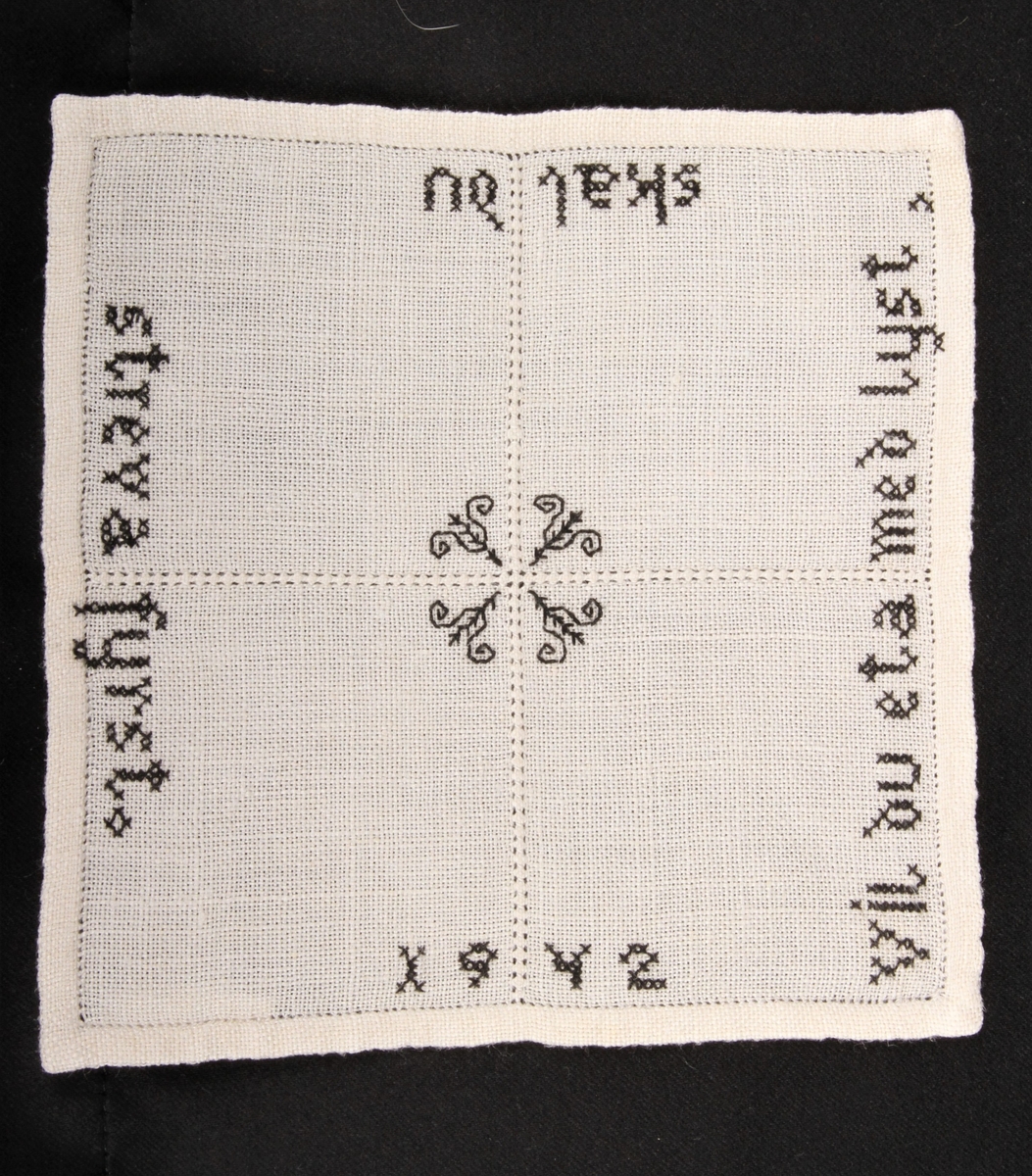 Vevd, naturfarge med svart dekor, brodert korsting, hullsøm, attersting. Border, tekst og blomster.