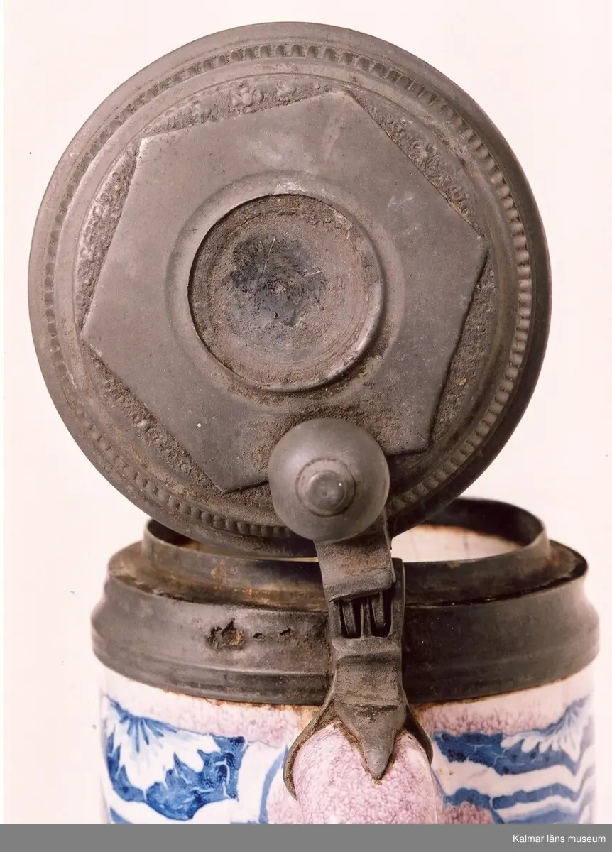 KLM 3004. Krus av fajans. Cylindrisk stop med handtag och tennlock. Ornering blåvita blommor och blad på violetta fält, inramademed kolonner och bågar. På lockets insida tre stämplar: C7 under krona (Christian VII), GG över hjortdjur med fana, stadsstämpel med krönt griphuvud. Tumgrepp: kula med krönknopp.