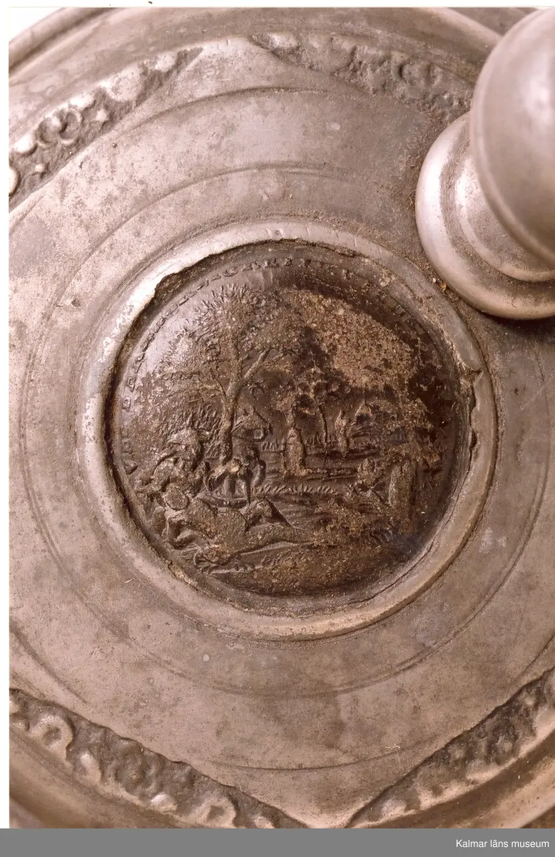 KLM 5725. Krus av fajans. Cylindrisk med grepe och tennlock, vari avbildning av en tysk medalj. På kruset ett landskap med hjort och hund, allt i blått, violett och gult. Tumgrepp: kula med gördel och krönknopp. Tennskodd botten. Tillverkat i Tyskland.