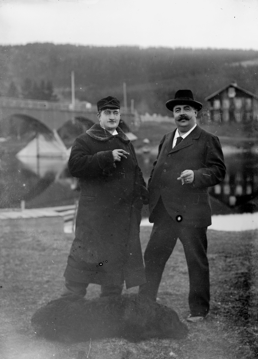 To menn og en hund foran bru over elva. Til venstre: Doktor (distriktslege i Trysil) Otto Mejlænder (1865-1944). Bildet er tatt på vestsida av Trysilelva, i nærheten av fotograf Engerdals hjem og atelier på Nybakk. Den gamle brua i Nybergsund sto fra 1879-1929. Huset i bakgrunnen er Folkvang, sentrum for varehandelen i Nybergsund fra 1870-årene (Trysilboka bind 6 s. 33) Doktor Mejlænder kom til Trysil som kommunelege i 1891. Mannen til høyre kan være Peder Grambo (1867-1927) eller Engebret Moe (1841-1911)