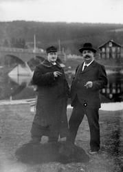 To menn og en hund foran bru over elva. Til venstre: Doktor 