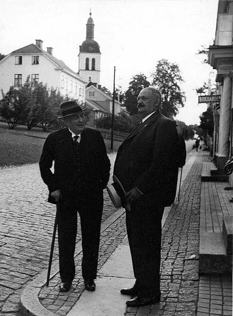 Två män står på trottoaren utanför Hallska gården i Gränna. Den äldre mannen är Sven Hedin. Han bär hatt och håller en käpp i höger hand. Till höger står en kraftigare och längre man som håller sin stråhatt i vänster hand, han är A E Bolling.