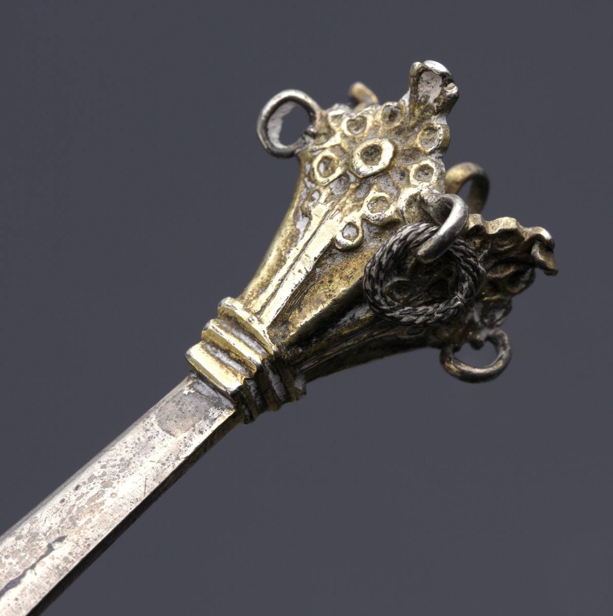 Brännvinssked av silver. 
S.k. kronsked med nästan runt skedblad, kort platt skaft, dekorerat med tvärräfflor längst ner. Skaftknopp i form av krona, 
sammansatt av fyra gotiska korsblommor. I kronans hörn hänger spiralvridna ringar (två saknas). 
Bladet har dekor i form av ett förgyllt kors med en graverad blomma i mitten. Även inre kanten av bladet är förgyllt. 
Stämplar och s.k. åldermansranka på skaftets baksida.

Inskrivet i huvudbok 1867.