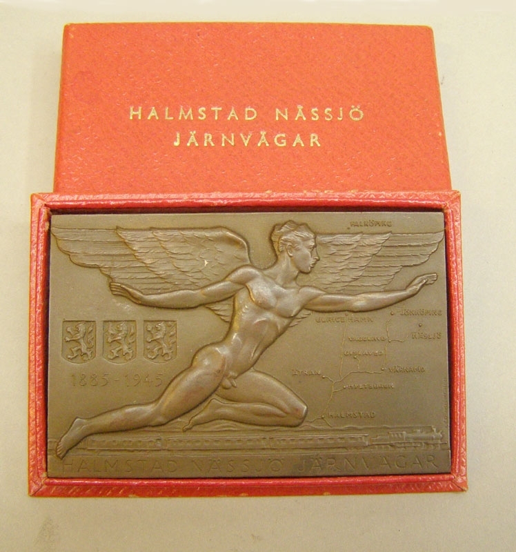 Rektangulär bronsplakett från Halmstad Nässjö Järnvägsaktiebolag.
På framsidan en bevingad mansfigur över en karta till höger, t v tre vapensköldar och årtalen 1885 - 1945. Nederst ett tåg draget av ett ånglok, samt texten "Halmstad Nässjö Järnvägar".