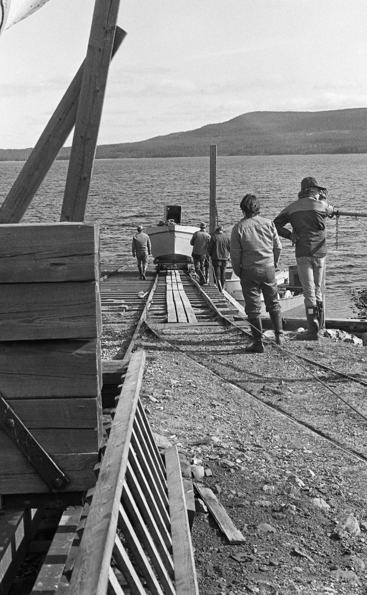 Utsetting av en av de tre varpebåtene som skulle brukes under tømmerfløtinga på Osensjøen i grenseområdet mellom Åmot og Trysil kommuner i Hedmark i 1984. Varpebåtene hadde 6-7 meter lange stålskrog, var drøyt 2 meter brede og hadde dieselmotor, som kunne drive både propellen, som sørget for framdrift, og en vinsj, som ble brukt ved oppsamling av løstømmer i små bommer. Varpebåtene ble vinterlagret i et eget båthus med bortimot 200 kvadratmeters grunnflate. Derfra gikk det en forholdsvis spinkel skinnegang med 110 centimeters sporbreddemot slippen, som også hadde en noe solidere skinnegang for den store og tunge slepebåten «Trysilknut», som ble vinterlagret på land, uten takoverbygg. Ved utsetting og opptak av varpebåtene og inspeksjonsbåten «Ener», som stod i båthuset vinterstid, ble det brukt ei skinnegående vogn som kunne vinsjes ned- eller oppover det skråplanet slippen var ved sjøsetting og opptak. Osensjøen er regulert, og nivåforskjellen mellom laveste og høyest tillatte vannstand er 6,6 meter. Dette fotografiet er tatt i vårsesongen, da sjøen var kraftig nedtappet, så avstanden ned til vannspeilet var ganske lang.

Den konstruksjonen vi ser litt av ytterkanten på til venstre i forgrunnen er den skinnegående vogna som slepebåten «Trysilknut» sto på i vinterhalvåret.