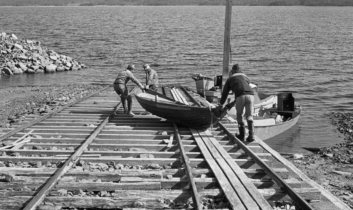 Utsetting av robåt som skulle brukes under tømmerfløtinga på Osensjøen i grensetraktene mellom Åmot og Trysil kommuner i Hedmark våren 1985. Fotografiet er tatt på Sørlistøa, der Glomma fellesfløtingsforening hadde kontor- og mannskapsbrakke, samt lagerhus til det materiellet som bare var i bruk i driftsperiodene om våren og på forsommeren. Den store slepebåten «Trysilknut» ble trukket på land på ei solid vogn på en trersporet skinnegang på slippen dette fotografiet er tatt fra. Parallelt med denne gikk det en noe spinklere skinnegang for den vogna som ble brukt når de mindre båtene skulle settes ut eller tas opp fra sjøen. Her brukte man imidlertid ikke den vogna som var lagd for de lettere farkostene. I stedet var det lagt bord mellom skinnene som robåtene kunne dras og skyves på uten at det slet for hardt på kjølsonen. Da dette fotografiet ble tatt kan det imidlertid virke som om karene forsøkte å flytte båten over i den delen av skinnegangen som ikke var bordlagt. Osensjøfløterne disponerte 5-6 slike drøyt 5 meter lange robåter, samt tre varpebåter med stålskrog. I tillegg hadde den lokale fløtersjefen en enspeksjonsbåt, «Ener», som også hadde stålskrog. De sistnevnte båtene lå fortøyd ved slippen, ba mannen som skjøv på akterenden av robåten, da dette fotografiet ble tatt.