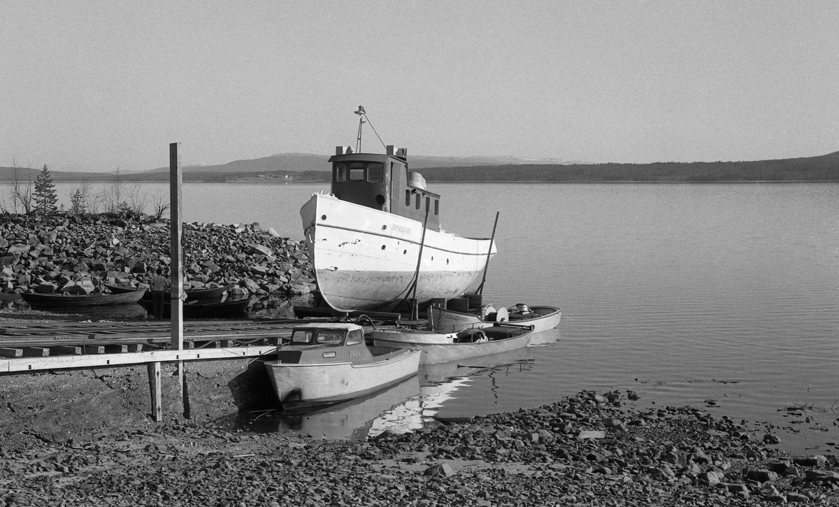 Sjøsetting av slepebåten Trysilknut fra Sørlistøa ved Osensjøen i mai 1984. Dette fartøyet ble trukket opp på slipp når fløtingssesongen, og der ble den stående vinteren over, inntil forberedelsene til neste fløtingssesong startet. På slippen sto Trysilknut på ei vogn, som kunne beveges opp og ned fra vannet på jernbaneskinner.  Avstanden mellom de ytterste skinnene var 360 centimeter. Da Trysilknut kom ut i vannet brukte fløterne varpebåten til å trekke den tunge slepebåten av vogna den hadde stått på gjennom vintersesongen. Dette fotografiet er tatt mens Trysilkut var på veg nedover slippen, men før båten nådde vannspeilet. Inspeksjonsbåten «Ener» og de to varpebåtene «Fløtarlars» og «Lykkjeknut» lå fortøyd ved sida av slippen. 

Osensjøen har et nedslagsfelt på 1 300 kvadratkilometer, hvorav om lag halvparten er bevokst med produktiv skog, og i dette området vokser det mye kvalitetsvirke.  Fra gammelt av var fløtinga her et samspill mellom strømdrag i sjøen, vinden og menneskelig muskelkraft.  Lenge ble stokkene samlet i flåter og forsøkt seilt mot Valmen og avløpselva Søndre Osa, noe som innebar vanskeligheter når det var vindstille eller motvind.  Utover på 1800-tallet begynte man å bruke spillflåter med gangspill, forankret i «dregger» (ankere) med lange trosser.  Denne formen for fløting la beslag på mye mannskap, og med stigende lønninger ble det en kostnadsfaktor tømmerkjøperne gjerne ville krympe.  I 1913 foreslo fløtingsdirektør Johs. Johannesen for styret i Christiania Tømmerdirektion (seinere Glomma fellesfløtingsforening) at organisasjonen skulle kjøpe en «slæpedamper» til fløtinga på denne innsjøen. Med denne investeringa antydet han at de årlige driftskostnadene kunne redusere driftskostnadene til snaut 40 prosent av hva de hadde vært. Styret gikk inn for saken – man ville få bygget en dampslepebåt til fløtinga på denne innsjøen.  Glommens mek. verksted i Fredrikstad fikk oppdraget.  De bygde tømmerslepebåten som en litt krympet utgave av D/S Storsjø, som var levert til fløtinga på Storsjøen i Rendalen ett år tidligere. Båten kostet drøyt 38 000 kroner.  Den ble transportert i seksjoner på jernbanen til Rena, og derfra på sleder til Osen vinteren 1914. Der ble seksjonene klinket sammen, slik at båten kunne sjøsettes når våren kom.  Trysilknut ble sjøsatt 2. mai 1914. Kaptein og maskinist hyret man i Fredrikstad-regionen. De første åra hadde båten base i og ved Valmen, men i slutten av 1930-åra bygde Glomma fellesfløtingsforening fløtingssenteret Sørlistøa, hvor det blant annet var slipp for Trysilknut, slik at båten kunne tas på land hver vinter. En «normal» tømmerbom for den dampdrevne slepebåten besto av 24 000 tømmerstokker og hadde et volum på om lag 4 000 kubikkmeter. Både mannskapet og ingeniørene i Glomma fellesfløtingsforening ønsket imidlertid å styrke trekkevnen, noe som ble realisert i 1957-58, da den 75 hestekrefters dampmaskinen ble demontert og erstattet av en 135 hestekrefters dieselmotor. Samtidig fikk båten nytt styrhus og nye lugarer. Dette fotografiet viser båten med det utseendet den fikk etter ombygginga. Det opprinnelig mørke skroget ble kvitmalt, riktignok med ei grønn linje. Styrhuset og byssa ble malt med en rødbrun farge, og den lyseblå skorsteinen fikk Glomma fellesfløtingsforenings initialer. Da det var klart at 1984 ville bli den siste sesongen med tømmerfløting på Osensjøen, tilbød Glomma fellesfløtingsforening Norsk Skogbruksmuseum å kjøpe Sørlistøa, båtene og det øvrige utstyret som lå igjen etter virksomheten. Museet aksepterte tilbudet, og startet arbeidet å bygge om Sørlistøa til museum.  Anlegget ble åpnet for publikum i 1989. Trysilknut var en del av attraksjonen.  Den ble sertifisert for mannskap og inntil 19-20 passasjerer, og gikk søndagsturer på sjøen i sommersesongen. Sjølve skroget var i bemerkelsesverdig god stand, men både oppdatering i tråd med nye sikkerhetsforskrifter og utbedring av fukt- og råteskader på interiøret måtte til.  Riksantikvaren bidro til finansiering, og Hardanger fartøyvernsenter var hovedansvarlige for det praktiske arbeidet, som ble utført i 2011-2012. I 2013 ble det elektriske anlegget utbedret, og i 2014 ble fartøyet malt med de fargene den fikk i 1958, og som det fortsatt hadde da dette fotografiet ble tatt.