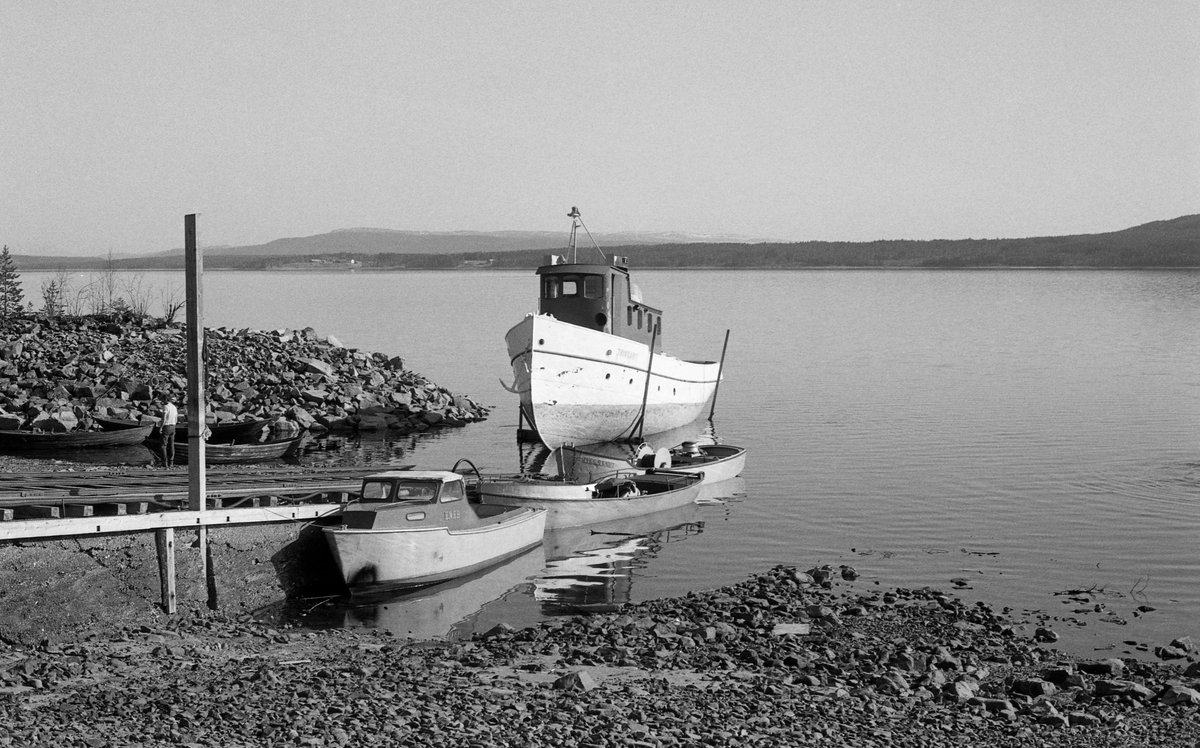 Sjøsetting av slepebåten Trysilknut fra Sørlistøa ved Osensjøen i mai 1984. Dette fartøyet ble trukket opp på slipp når fløtingssesongen, og der ble den stående vinteren over, inntil forberedelsene til neste fløtingssesong startet. På slippen sto Trysilknut på ei vogn, som kunne beveges opp og ned fra vannet på jernbaneskinner.  Avstanden mellom de ytterste skinnene var 360 centimeter. Da Trysilknut kom ut i vannet brukte fløterne varpebåten til å trekke den tunge slepebåten av vogna den hadde stått på gjennom vintersesongen. Dette fotografiet er tatt mens Trysilkut var på veg nedover slippen, men før båten nådde vannspeilet. Inspeksjonsbåten «Ener» og de to varpebåtene «Fløtarlars» og «Lykkjeknut» lå fortøyd ved sida av slippen. 

Osensjøen har et nedslagsfelt på 1 300 kvadratkilometer, hvorav om lag halvparten er bevokst med produktiv skog, og i dette området vokser det mye kvalitetsvirke.  Fra gammelt av var fløtinga her et samspill mellom strømdrag i sjøen, vinden og menneskelig muskelkraft.  Lenge ble stokkene samlet i flåter og forsøkt seilt mot Valmen og avløpselva Søndre Osa, noe som innebar vanskeligheter når det var vindstille eller motvind.  Utover på 1800-tallet begynte man å bruke spillflåter med gangspill, forankret i «dregger» (ankere) med lange trosser.  Denne formen for fløting la beslag på mye mannskap, og med stigende lønninger ble det en kostnadsfaktor tømmerkjøperne gjerne ville krympe.  I 1913 foreslo fløtingsdirektør Johs. Johannesen for styret i Christiania Tømmerdirektion (seinere Glomma fellesfløtingsforening) at organisasjonen skulle kjøpe en «slæpedamper» til fløtinga på denne innsjøen. Med denne investeringa antydet han at de årlige driftskostnadene kunne redusere driftskostnadene til snaut 40 prosent av hva de hadde vært. Styret gikk inn for saken – man ville få bygget en dampslepebåt til fløtinga på denne innsjøen.  Glommens mek. verksted i Fredrikstad fikk oppdraget.  De bygde tømmerslepebåten som en litt krympet utgave av D/S Storsjø, som var levert til fløtinga på Storsjøen i Rendalen ett år tidligere. Båten kostet drøyt 38 000 kroner.  Den ble transportert i seksjoner på jernbanen til Rena, og derfra på sleder til Osen vinteren 1914. Der ble seksjonene klinket sammen, slik at båten kunne sjøsettes når våren kom.  Trysilknut ble sjøsatt 2. mai 1914. Kaptein og maskinist hyret man i Fredrikstad-regionen. De første åra hadde båten base i og ved Valmen, men i slutten av 1930-åra bygde Glomma fellesfløtingsforening fløtingssenteret Sørlistøa, hvor det blant annet var slipp for Trysilknut, slik at båten kunne tas på land hver vinter. En «normal» tømmerbom for den dampdrevne slepebåten besto av 24 000 tømmerstokker og hadde et volum på om lag 4 000 kubikkmeter. Både mannskapet og ingeniørene i Glomma fellesfløtingsforening ønsket imidlertid å styrke trekkevnen, noe som ble realisert i 1957-58, da den 75 hestekrefters dampmaskinen ble demontert og erstattet av en 135 hestekrefters dieselmotor. Samtidig fikk båten nytt styrhus og nye lugarer. Dette fotografiet viser båten med det utseendet den fikk etter ombygginga. Det opprinnelig mørke skroget ble kvitmalt, riktignok med ei grønn linje. Styrhuset og byssa ble malt med en rødbrun farge, og den lyseblå skorsteinen fikk Glomma fellesfløtingsforenings initialer. Da det var klart at 1984 ville bli den siste sesongen med tømmerfløting på Osensjøen, tilbød Glomma fellesfløtingsforening Norsk Skogbruksmuseum å kjøpe Sørlistøa, båtene og det øvrige utstyret som lå igjen etter virksomheten. Museet aksepterte tilbudet, og startet arbeidet å bygge om Sørlistøa til museum.  Anlegget ble åpnet for publikum i 1989. Trysilknut var en del av attraksjonen.  Den ble sertifisert for mannskap og inntil 19-20 passasjerer, og gikk søndagsturer på sjøen i sommersesongen. Sjølve skroget var i bemerkelsesverdig god stand, men både oppdatering i tråd med nye sikkerhetsforskrifter og utbedring av fukt- og råteskader på interiøret måtte til.  Riksantikvaren bidro til finansiering, og Hardanger fartøyvernsenter var hovedansvarlige for det praktiske arbeidet, som ble utført i 2011-2012. I 2013 ble det elektriske anlegget utbedret, og i 2014 ble fartøyet malt med de fargene den fikk i 1958, og som det fortsatt hadde da dette fotografiet ble tatt.