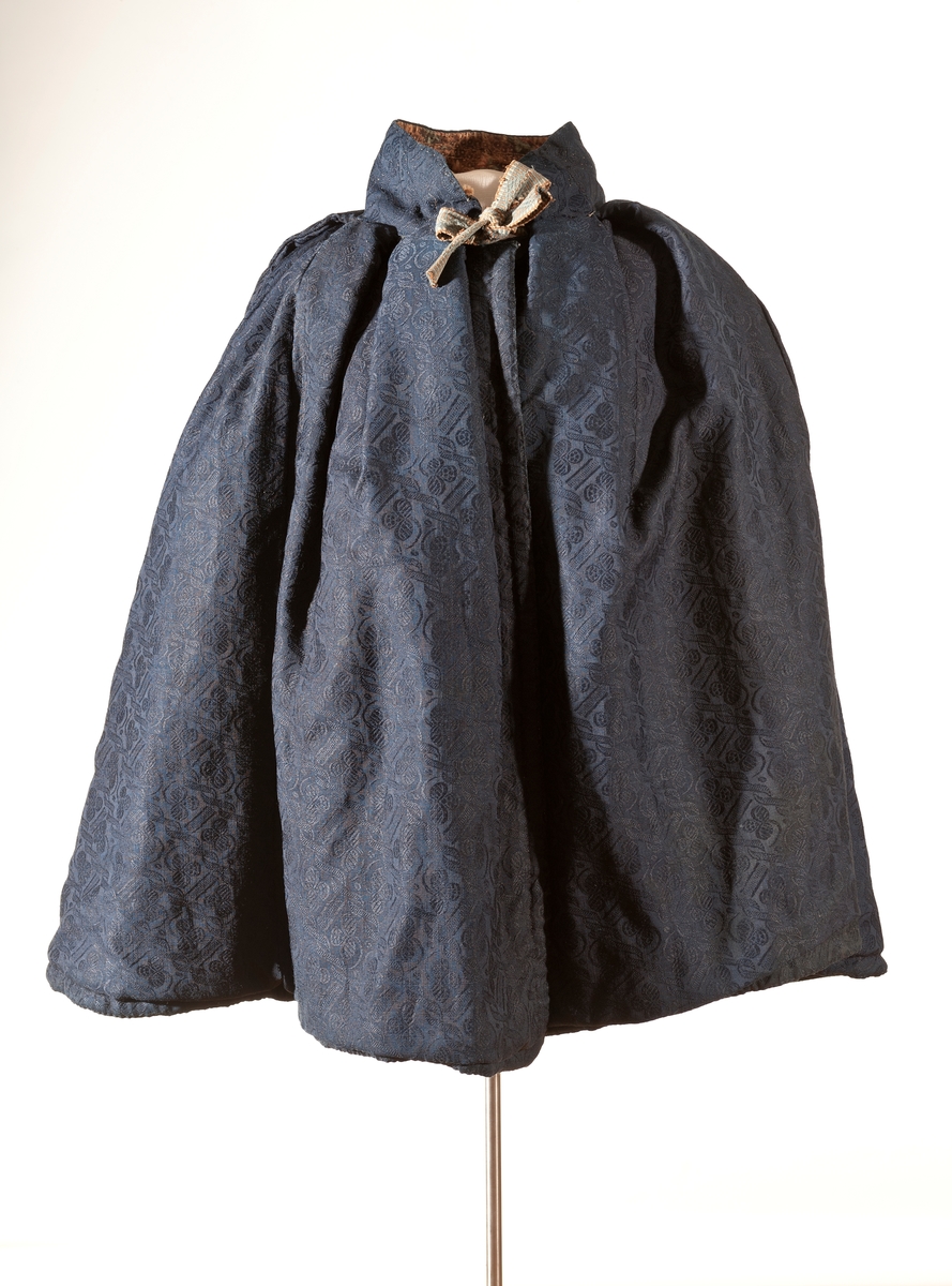 Mørk blå kep. Ytterstoff i everlasting. Fôr i hjemmevevet ullstoff i kypert. Halslinning er fôret med kattun (mørk brunt med blomster i rosa/blått). Kepen lukkes med ripsvevd knytebånd i halsen. 