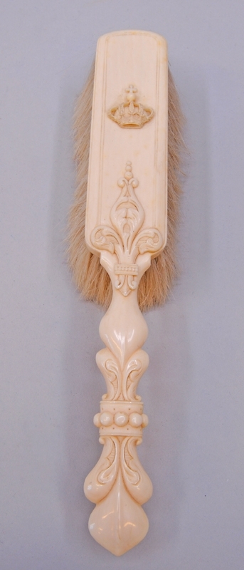 Gräddfärgad borste med handtag av elfenben och borst av animaliskt material, troligtvis gethår. Handtaget är format efter dess ornament som liknar akantusblad, mitt på handtaget finns pärlstavsdekor. Handtagets ornamentering fortsätter i relief en bit upp på borstens rygg. Ryggen är rektangulär och på mitten finns en kungakrona i relief.
