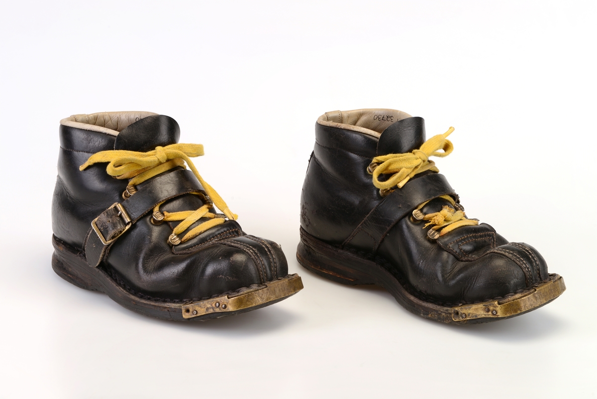 Et par dameskisko i svart skinn i størrelse 37 fra Falk Skofabrikk. Skoene er randsydde og har gummisåler. Foran er de firkantet på form og det er metallbeslag (messing?) på sålen for feste i skibinding. I front er det 4 par hekter for snøring. Under snøringen er det en enkel skinntunge. Skoene er snøret med gule skolisser. Over snøringen er det en skinnreim som festes i en spenne på utsiden av skoen. De er foret med gråhvitt skinn og tekstil med påskrift. Kanten langs åpningen er polstret. Innersålen er av brunt skinn. På området til hælen er det forsetrket med lyst skinn. I skoene ligger det løse ullsåler. Undersiden av yttersålen har rutemønster. Bakpå hælen er det fure for feste til binding.