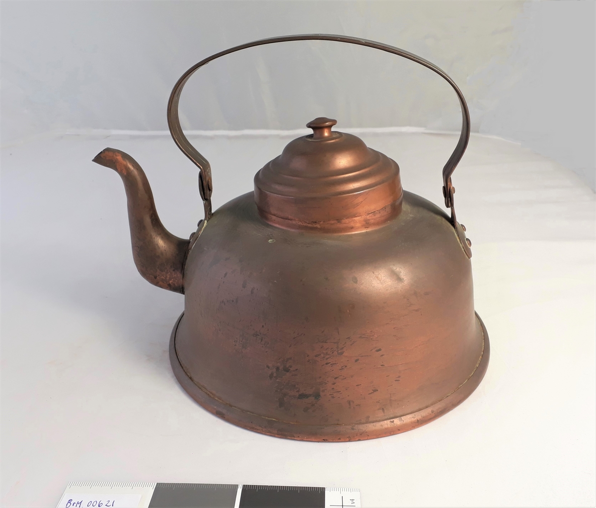 Kaffekjele i kobber med lokk. Sirkulær med innsnevring mot lokket og utvidelse mot en bredere fotkant. Hankfestet ved tuten og på motsatt side.
