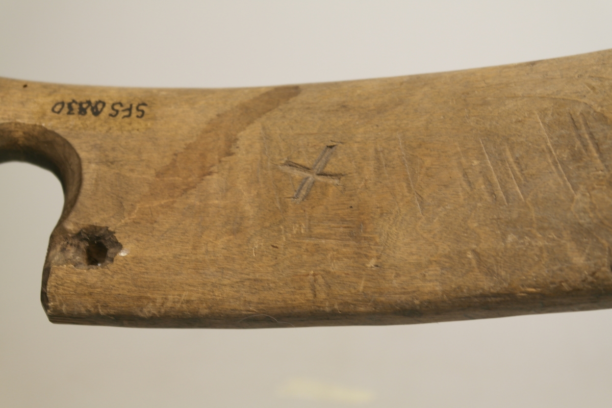 Sauestokk i tre. Stokken er buet og har en rund utsparing på midten samt to små hull på hver side av utsparingen. På den ene siden er det inngravert ett kryss samt et håndskrevet personnavn. En merkelapp med håndskrevet tekst har vært festet til stokken med hyssing. Stokken ble brukt for å hindre sau å hoppe over gjerde o.l. ved å feste den til sauens ben.