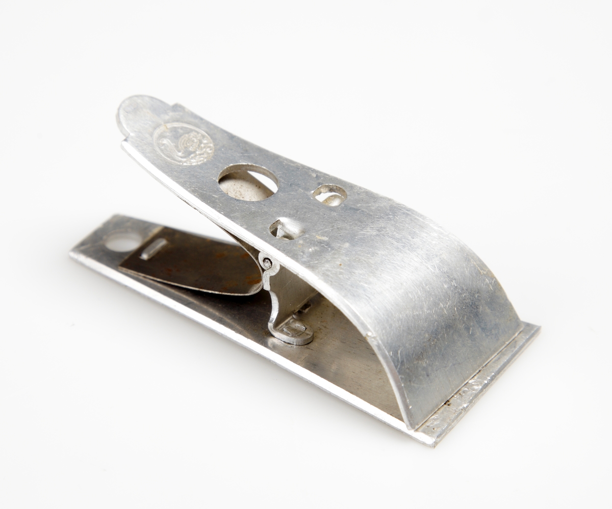 Pappersklämma i bockad aluminium försedd med klämfjäder. Översidan försedd med en symbol i form av en svan inom cirkel.
