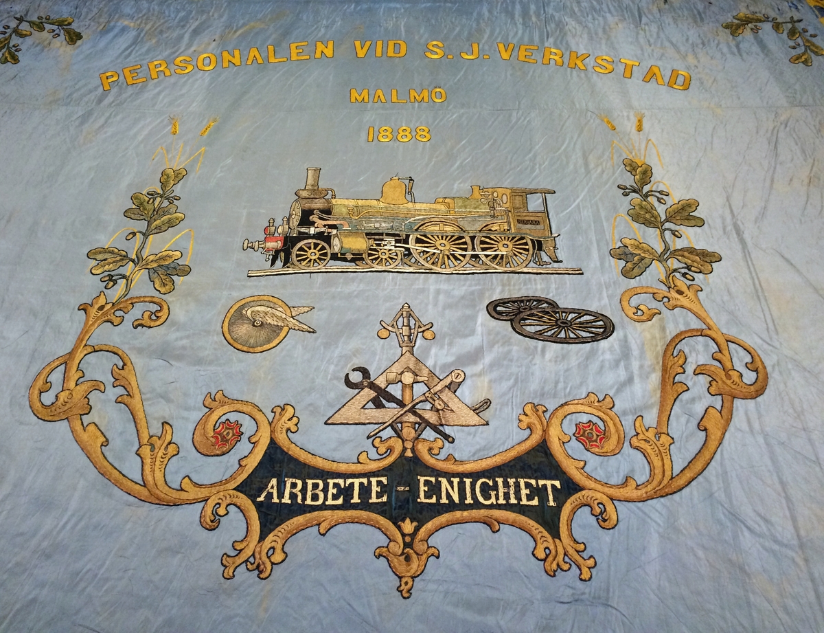 Rektangulär fackföreningsfana av blått broderat siden, för en järnvägsmannaförening i Malmö.
I mitten syns ångloket SJ Cb 339 Hervara, under detta ett bevingat hjul, en hjulaxel, samt ett arrangemang av verktyg och växtrankor.
Text i gult : "Personalen vid S.J. verkstad Malmö 1888 Arbete Enighet". Fanan är broderad i guld, svart och bruna nyanser med ett lok på mitten och eklöv i hörnen. Broderierna är utförda i stjälksöm och schattérsöm, vissa partier är även applicerade. Fanan har blågula fransar i kanterna.

Fanan är mycket blekt på den sidan som har legat uppåt i monter. Se sista bilden för jämförelse med undersidan.
