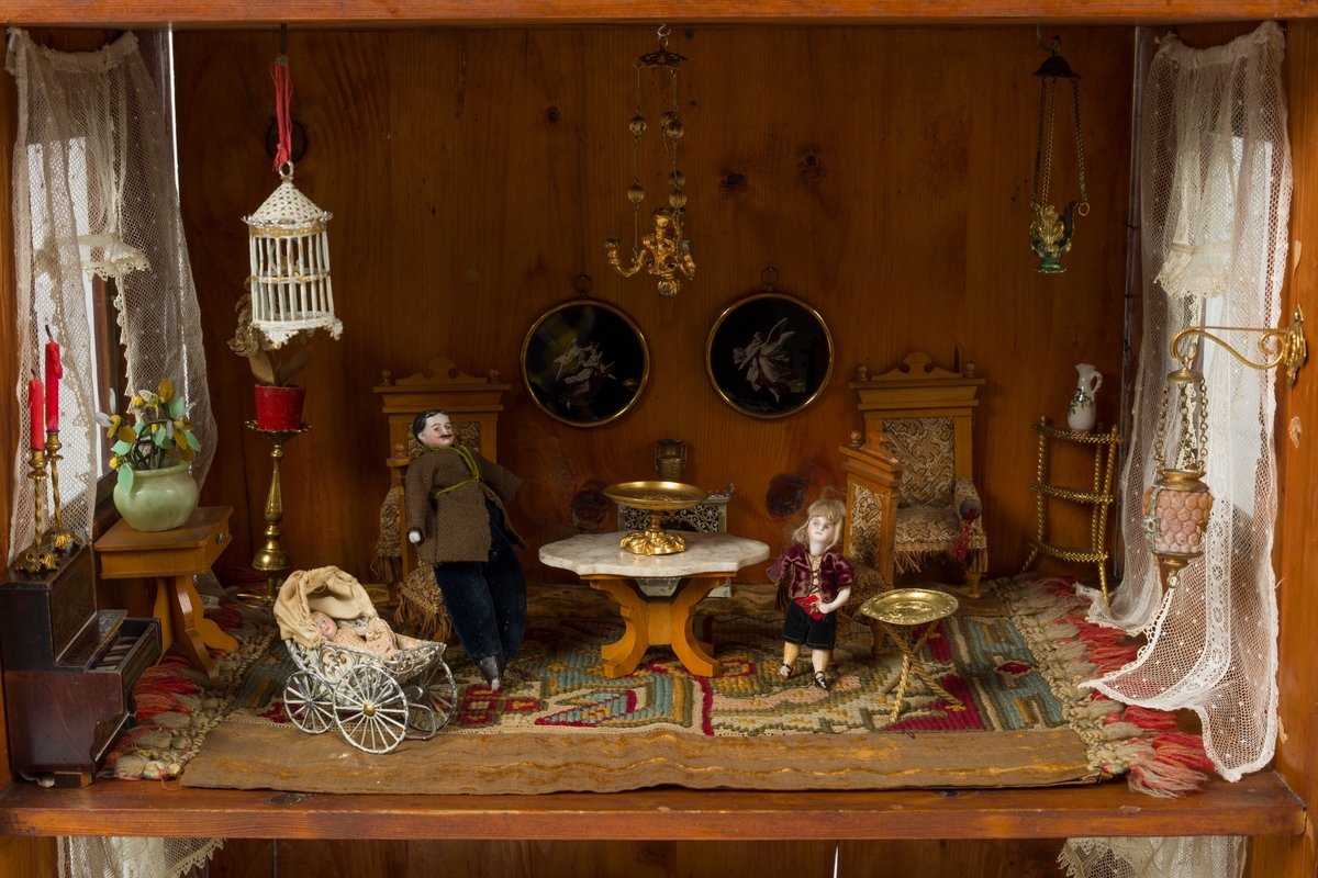 Dukkehus i tre. Huset har 3 etasjer og et spisstak. Hver etasje har 2 vinduer med gardiner. Overflaten er beiset og lakkert.