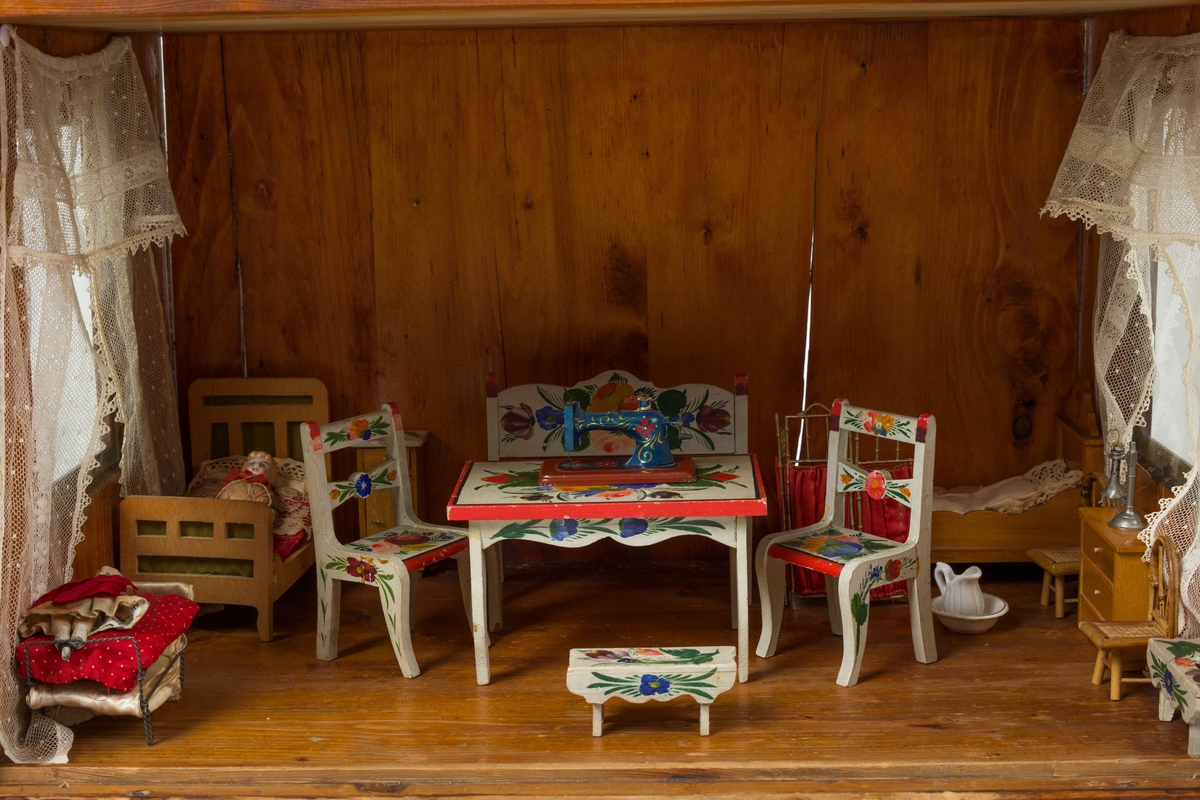 Dukkehus i tre. Huset har 3 etasjer og et spisstak. Hver etasje har 2 vinduer med gardiner. Overflaten er beiset og lakkert.