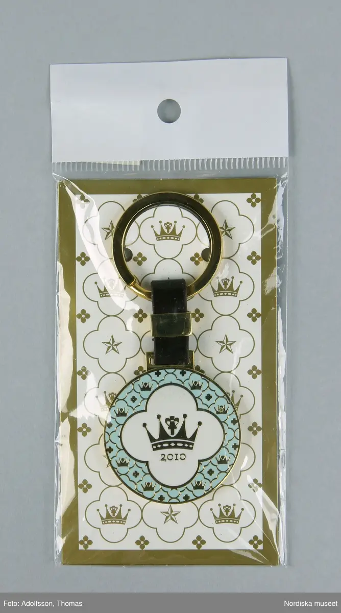 Nyckelring, i originalförpackning av transparent plast och papper. Nyckelringen består av en öppningsbar ring, en medalj med Den Officiella Bröllopsseriens logotyp av metall. Ringen och medaljen är sammanfogade med en rem av konstläder som hålls samman av en metallklipps. Ringen är fäst i en pappersbakgrund i förpackningen. På förpackningen en etikett med streckkod och artikelnummer. Ingår i "Den officiella Bröllopsserien". Se ref.materal ”Den Officiella Bröllopsserien” och www.upside.se

På nyckelringen: "2010", på förpackningen: "20102010 Den Officiella bröllopsserien The official Wedding Series Del av intäkterna går till Kronprinsessparets  Bröllopsstiftelse vars syfte är att motverka utanförskap och främja god hälsa bland barn och ungdomar i Sverige.","Part of the revenue is donated to the Crown Prinsess Couple's Wedding Foundation, which aims to prevent social isolation and promote good health among children and youths in Sweden", " UPSIDE AB www.upside.se +468109650