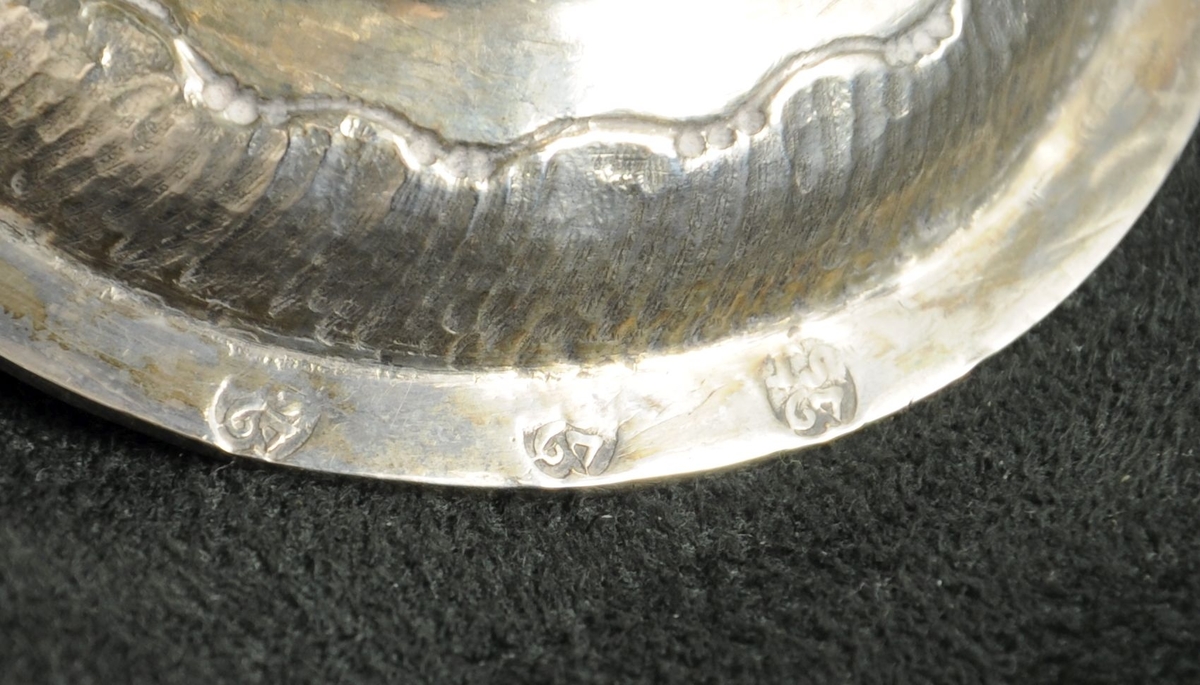 Beger av sølv. Begeret har stett og rund sokkel. På klokken er det to border med dekorative punslinger, mellom bordene er det drevet dekor av blomster. Siselert bølgedeker på sokkel.