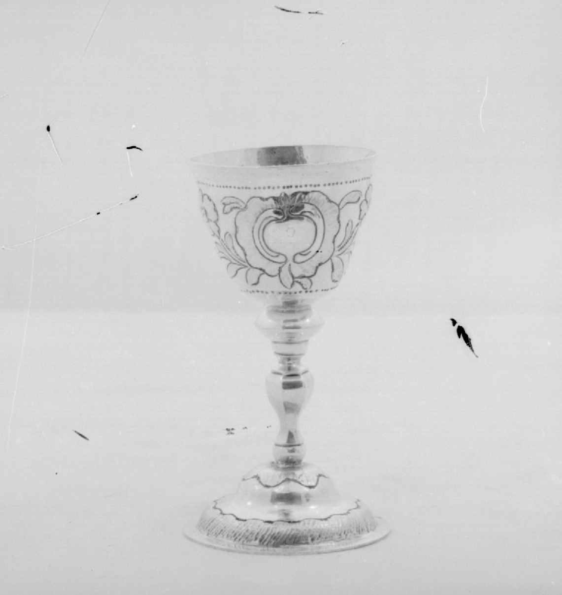 Beger av sølv. Begeret har stett og rund sokkel. På klokken er det to border med dekorative punslinger, mellom bordene er det drevet dekor av blomster. Siselert bølgedeker på sokkel.