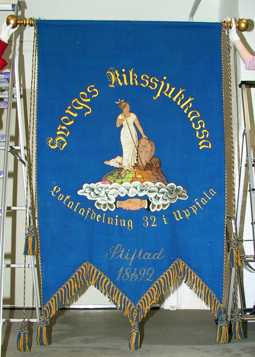 Av flaggblått ylle. Broderat med silke i schattérsöm i ett flertal färger. Moder Svea stående på jordklotet och text: Sveriges Rikssjukkassa Lokalavdelning 32 i Uppsala Stiftad 18 1/12 92. Kantat med blågula snoddar och tofsar. Fodrat med gult ylle. Trästång med svarvade knoppar. Bärstång av trä. Bärbälte av svart läder. Svart sorgflor längd ca 140 cm.