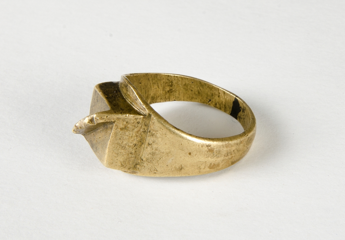 Fingerring av metall, sannolikt mässing. Lödning i "bakkant". Dekoren är en kvadrat med en upphöjd "kam" med fyra taggar. En trolig datering är historisk tid.