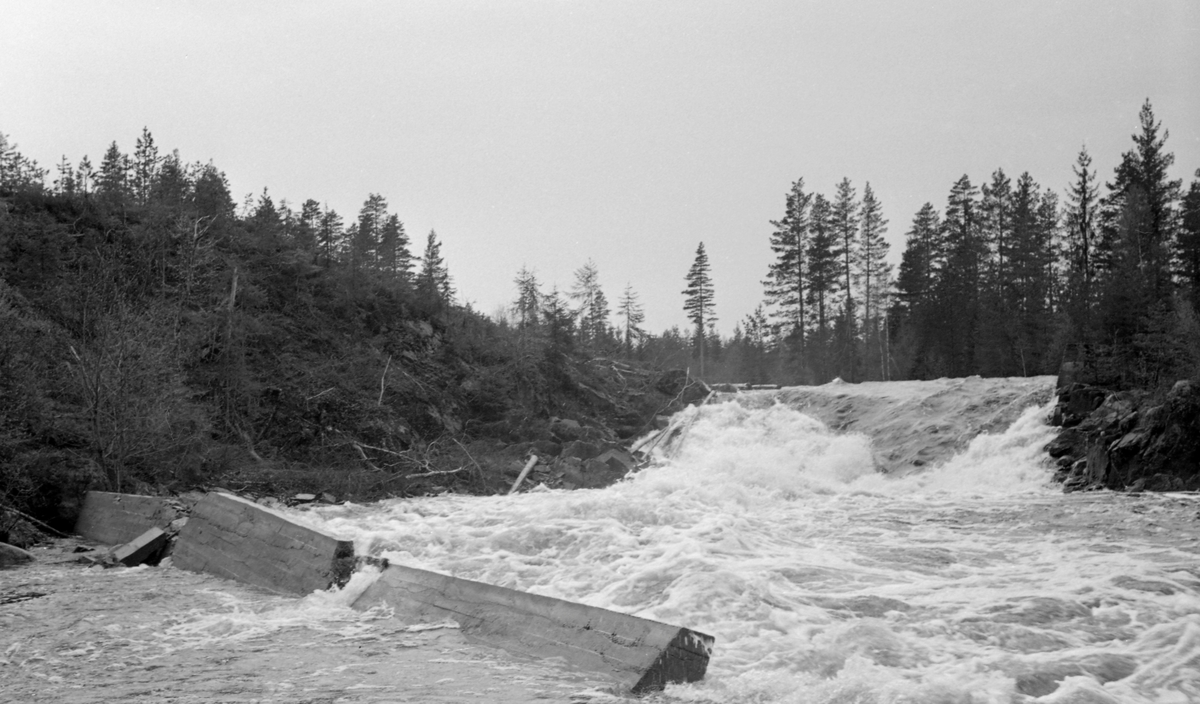 Betongskådam ved Brattfossen i Trautåa i Nord-Odal, fotografert våren 1954, året etter at denne dammen ble støpt. Skådammen var en mur, et skjerm som skulle hindre at tømmeret drev på land når vannføringa var høy, noe som ville ha ført til at stokkene etterpå måtte bæres tilbake til det strømmende vannet i elvløpet. Da dette fotografiet ble tatt var den cirka ett år gamle skådammen knekt i tre deler, og hadgde veltet inn i elveløpret. På grunn av forskyvninger mellom bruddstykkene var muren forvandlet fra en skjerm som skulle avvise tømmeret til et stengsel som fanget stokkene, og dermed et problem for fløterne, Om årsakene til at denne stokken fikk så kort levetid noterte fløtingsfunksjonæren som arkiverte dette fotografiet: «Dårlig blanding og elendig prosentsten».