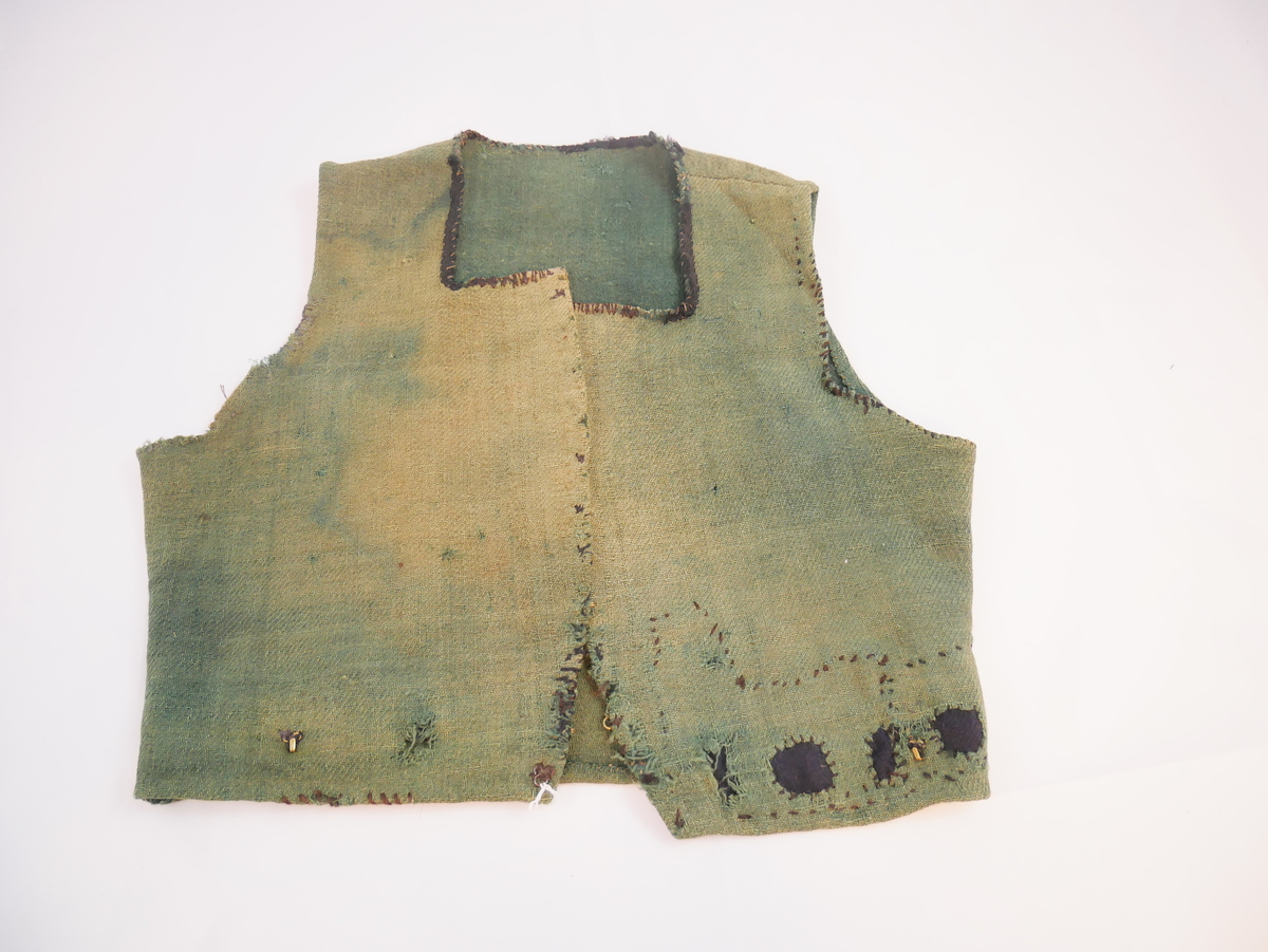 Livstykke som er overdel til kvinnedrakt. Livstykke i grønn ull med firskoren halsopning. Skal festast heilt ihop framme med 11 sett hekter og hemper. Hekter og hemper rundt livet til feste for skjørt. Rundt ermeopningane har det vore enkel fald, seinare er dette stadvis forsterka med overkast av grov tråd. Det same er tilfellet rundt halsopningen der det opphavleg har vore svart ullband. Livstykket er reparert med svarte og blå bøter i nedkant. Framkanten på livet er runda for å lage plass til bysten.